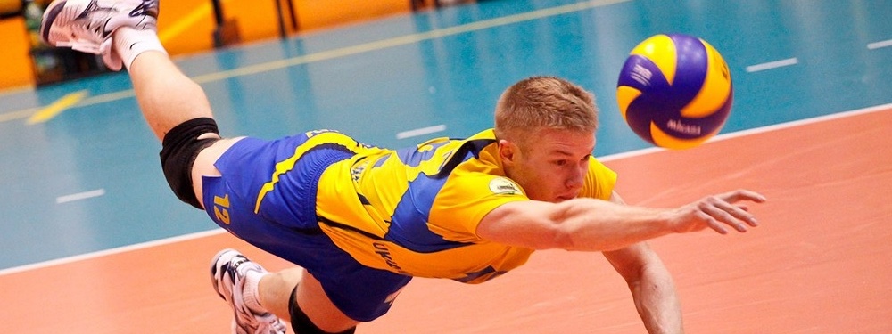 A guy leaping to dig a volleyball about to fall on the ground.